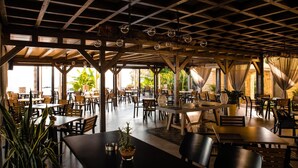 Un comedor interior con mesas y sillas de madera, ventanas amplias y plantas en macetas.
