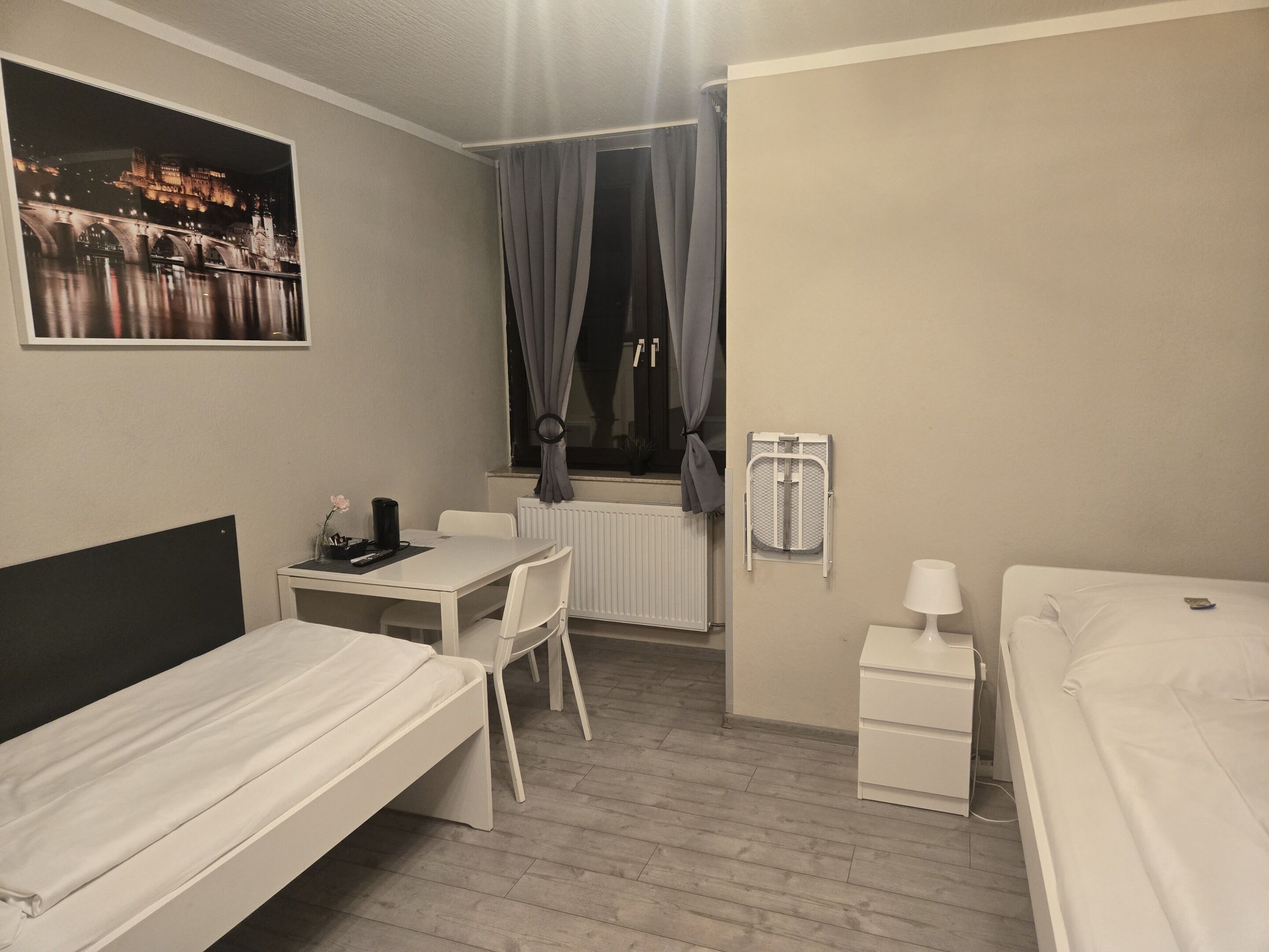 A compact hotel room with a bed, a small desk, a chair, a radiator, and a window with curtains.