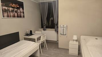A compact hotel room with a bed, a small desk, a chair, a radiator, and a window with curtains.