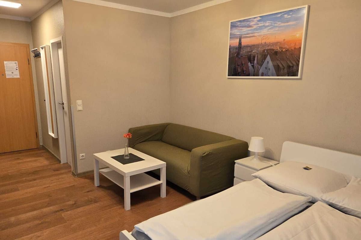 Ein Hotelzimmer mit einem grünen Sofa, zwei Einzelbetten, einem kleinen weißen Tisch mit einer Vase, einer Lampe und einem Stadtbild an der Wand.