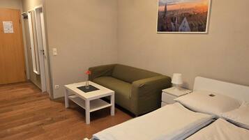 A hotel room with a green sofa, two single beds, a small white table with a vase, a lamp, and a painting of a cityscape on the wall.