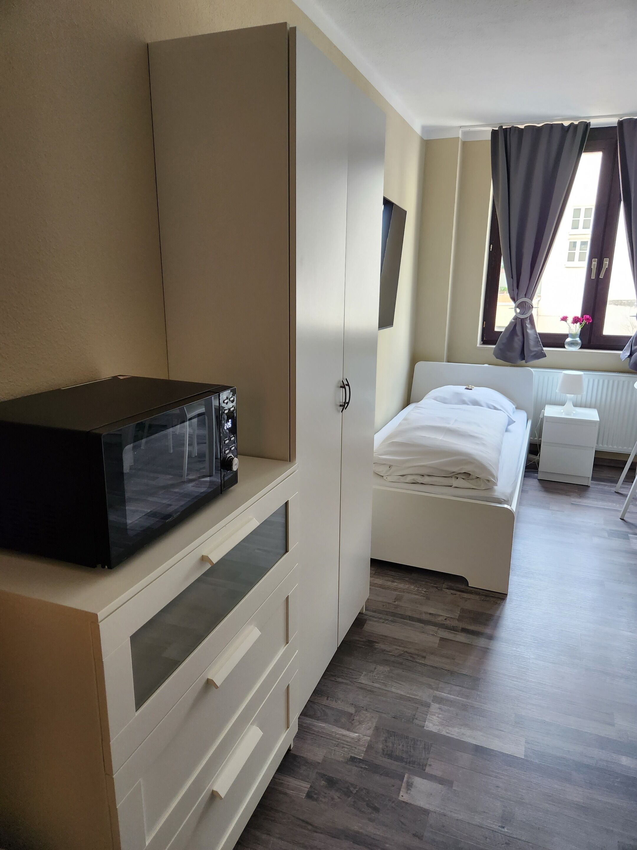 Ein Schlafzimmer mit einem Bett, einem weißen Kleiderschrank, einem Nachttisch, einer Lampe und einem Fenster mit Vorhängen.