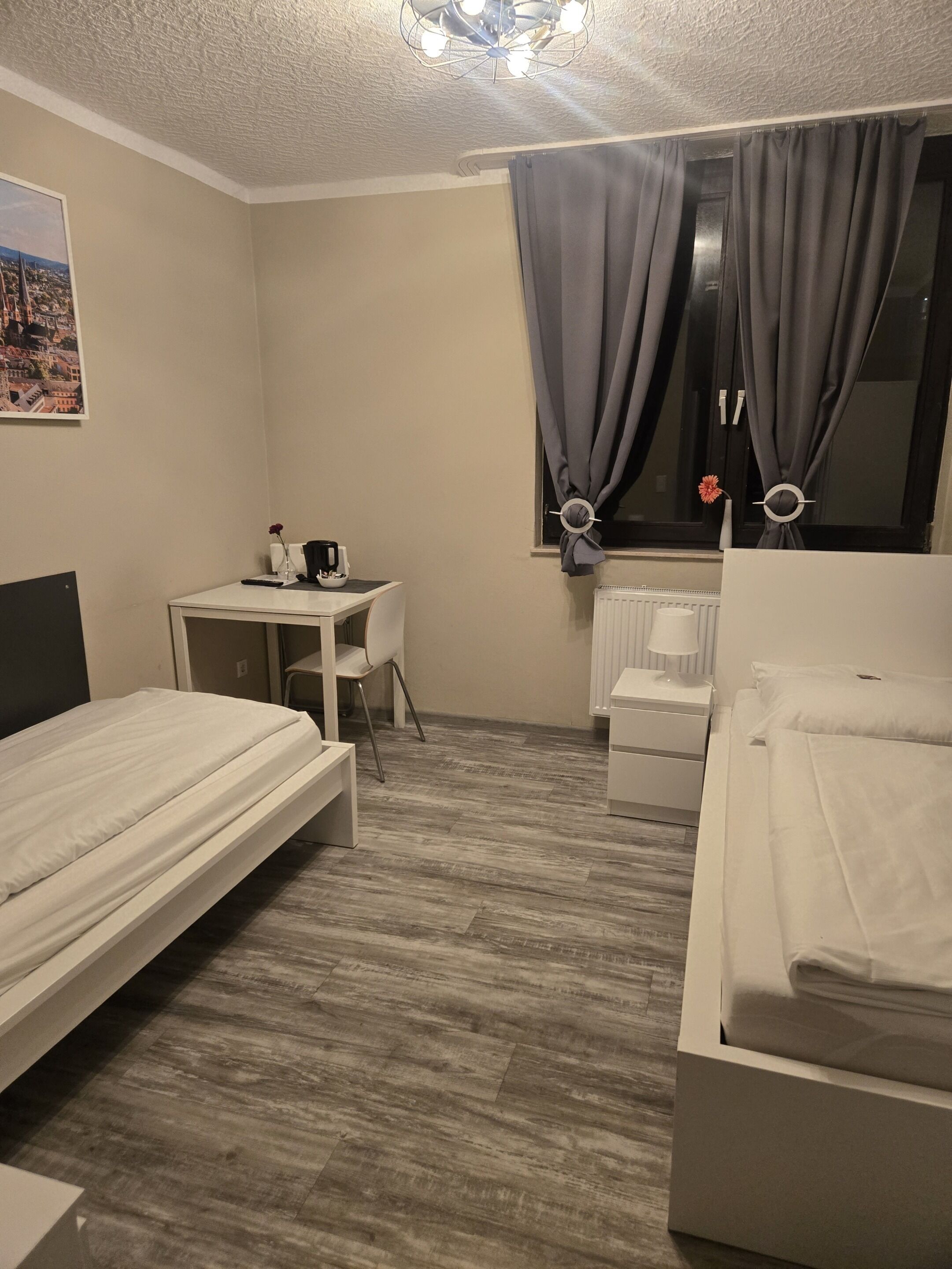 A hotel room with two single beds, a small desk with a chair, a radiator, and a window with curtains.