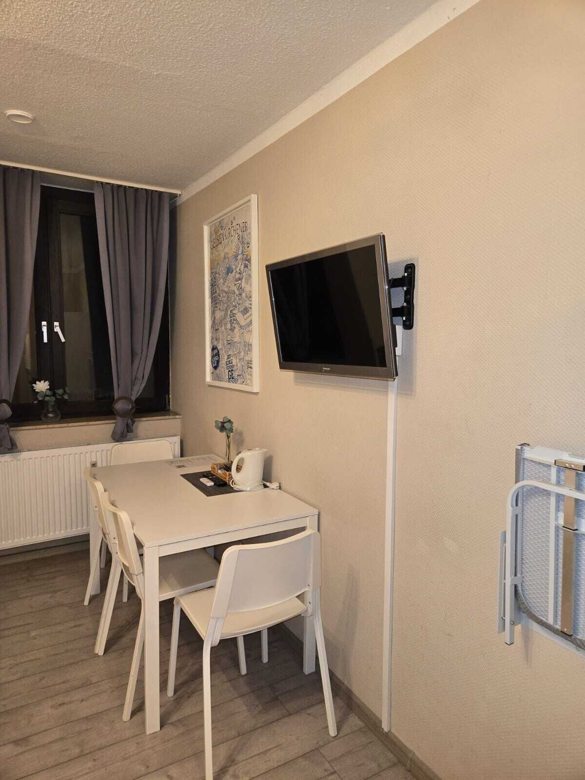 Una habitación pequeña y bien iluminada, con una mesa y sillas blancas, un televisor montado, un radiador y una ventana con cortinas.