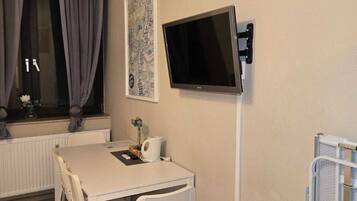 Une petite pièce bien éclairée, avec une table et des chaises blanches, un téléviseur fixé au mur, un radiateur et une fenêtre avec des rideaux.