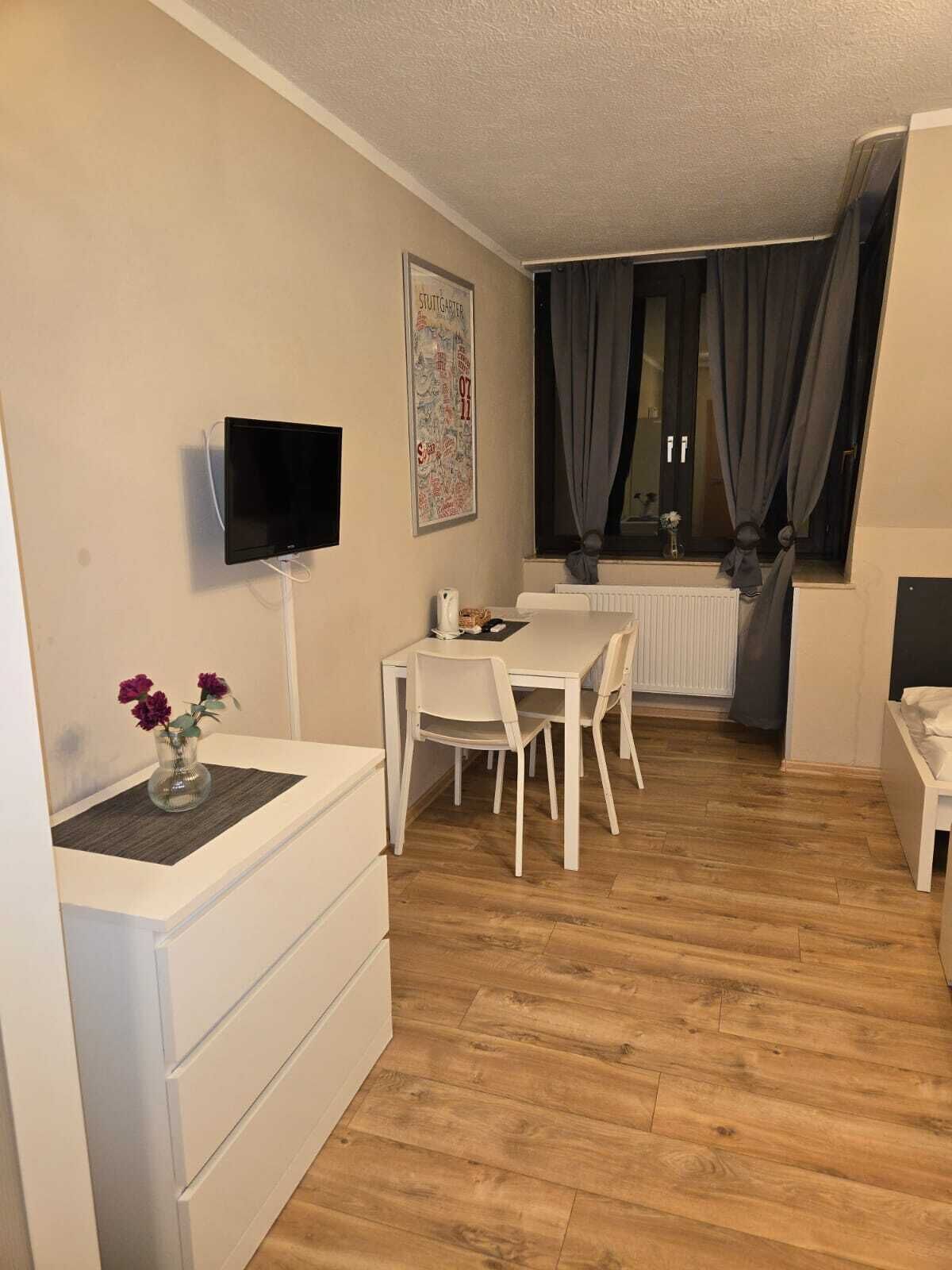 Ein kleiner, gut beleuchteter Raum mit einem weißen Tisch und Stühlen, einem flachbildfernseher an der Wand, einer Heizung und einem Fenster mit Vorhängen.