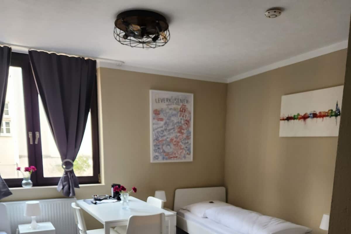 Ein Zimmer mit einem Bett, einem Schreibtisch und einem Stuhl. Es gibt ein Fenster mit Vorhängen, eine Heizung und ein Poster an der Wand.