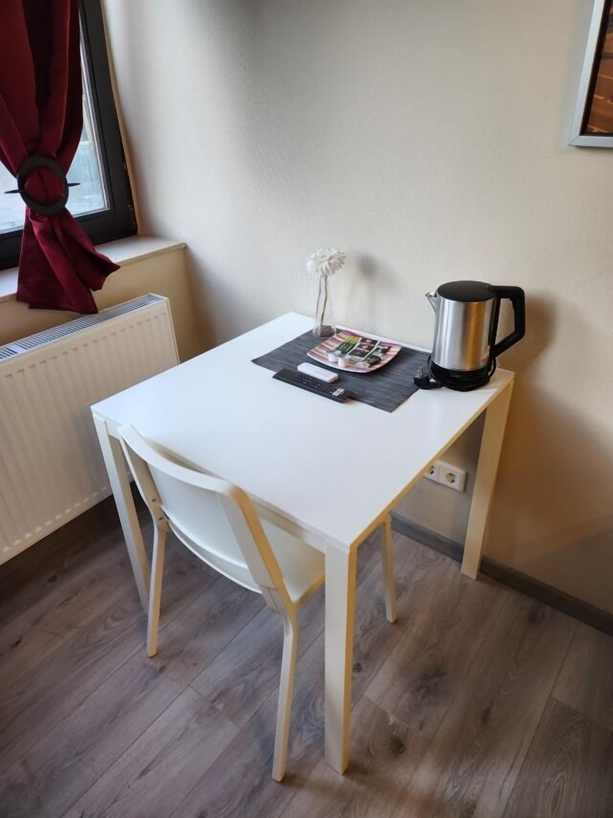 Uma pequena mesa branca com uma revista, uma chaleira preta e um vaso branco com uma única flor.
