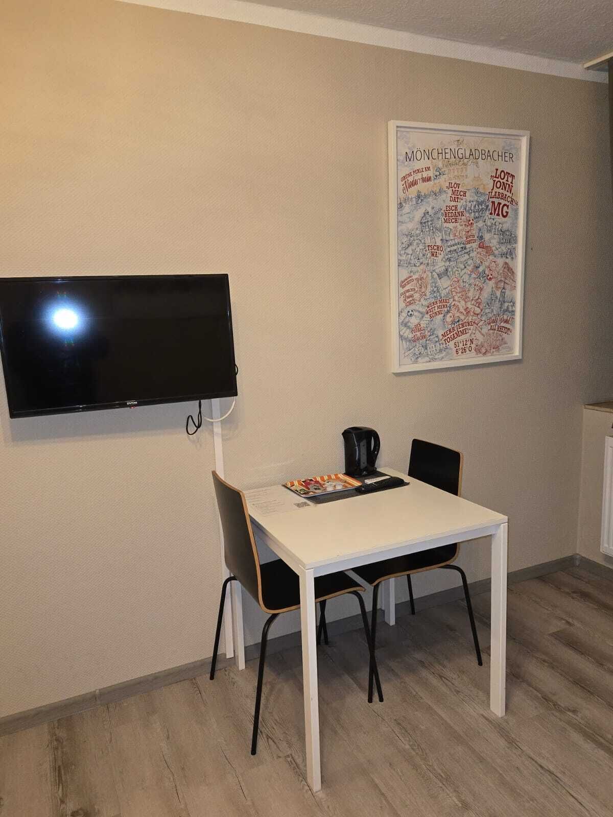 Una zona de menjador petita amb una taula blanca, dues cadires negres, una cafetera negra i un televisor de pantalla plana fixat a la paret.