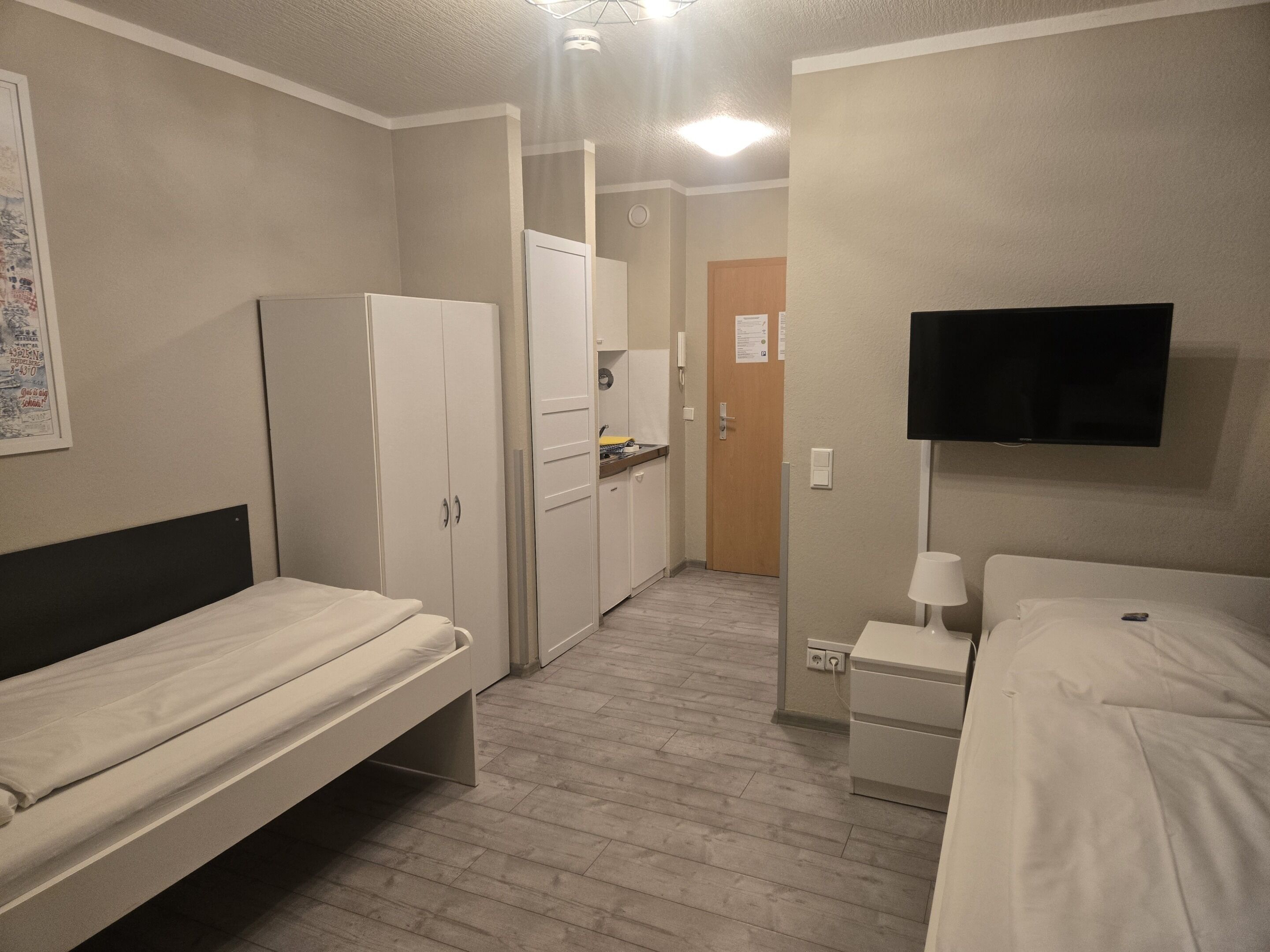 Una camera d'albergo con due letti singoli, un angolo cottura, una TV fissata al muro e un ingresso che conduce in un'altra stanza.