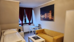 Een compacte hotelkamer met een bed, een bank, een kleine tafel, een raam met gordijnen, een radiator en een schilderij van een historisch gebouw.