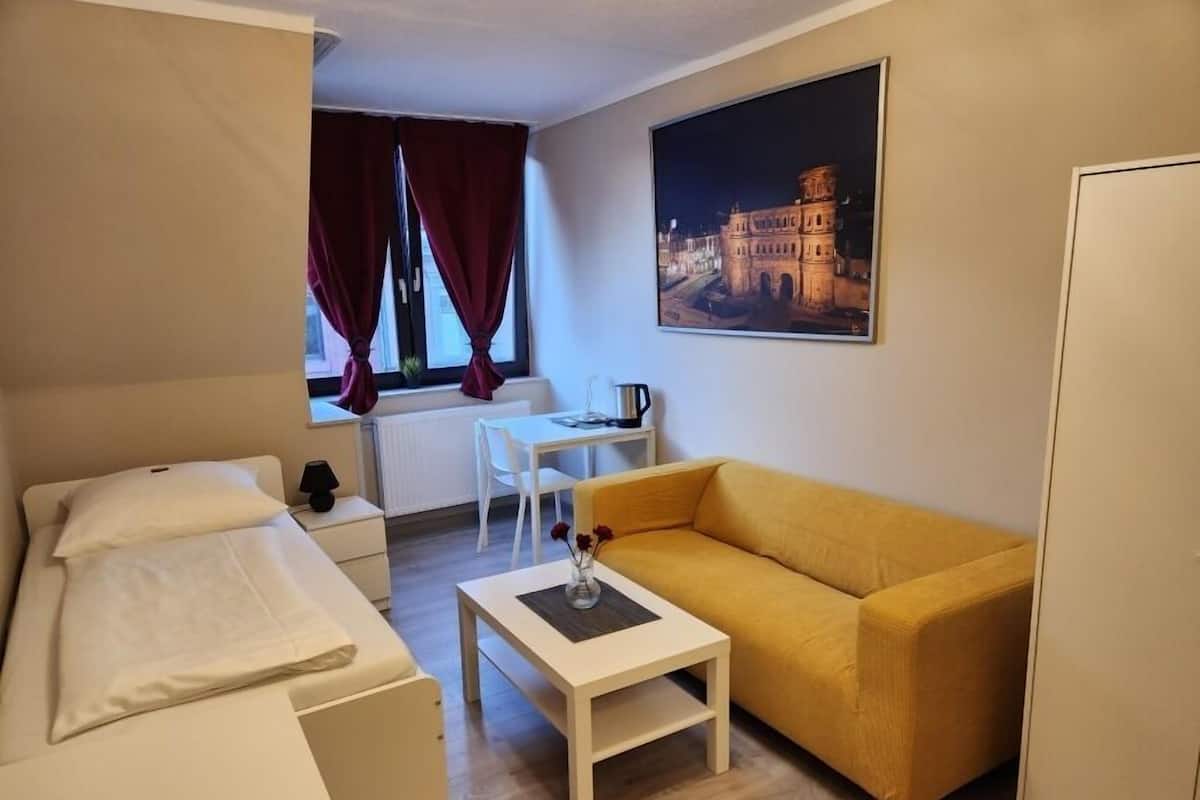 Ein kleines Hotelzimmer mit Bett, Sofa, einem kleinen Tisch, einem Fenster mit Vorhängen, einem Heizkörper und einem Gemälde eines historischen Gebäudes.