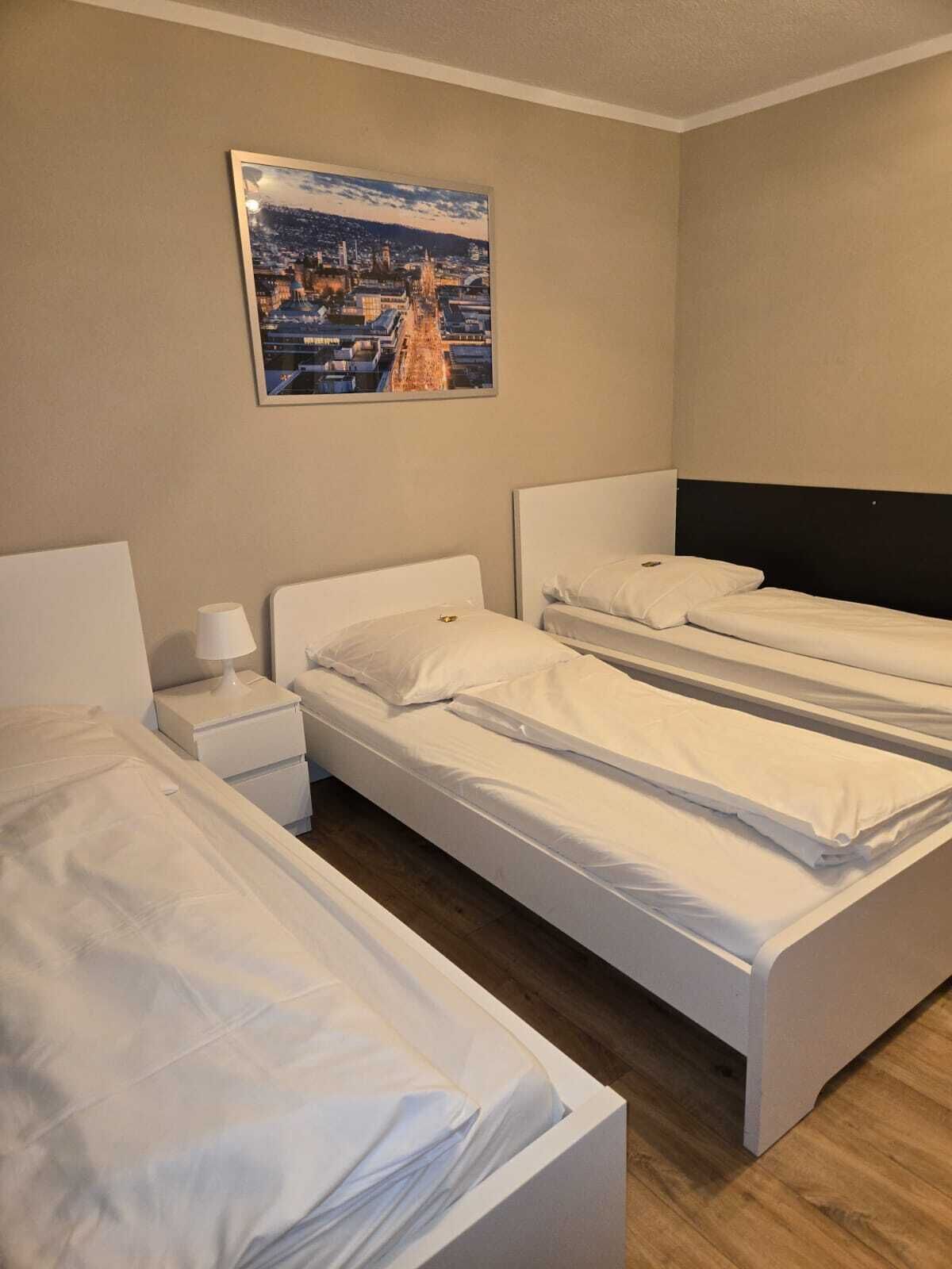 A hotel room with three single beds, a nightstand with a lamp, and a framed cityscape picture on the wall.