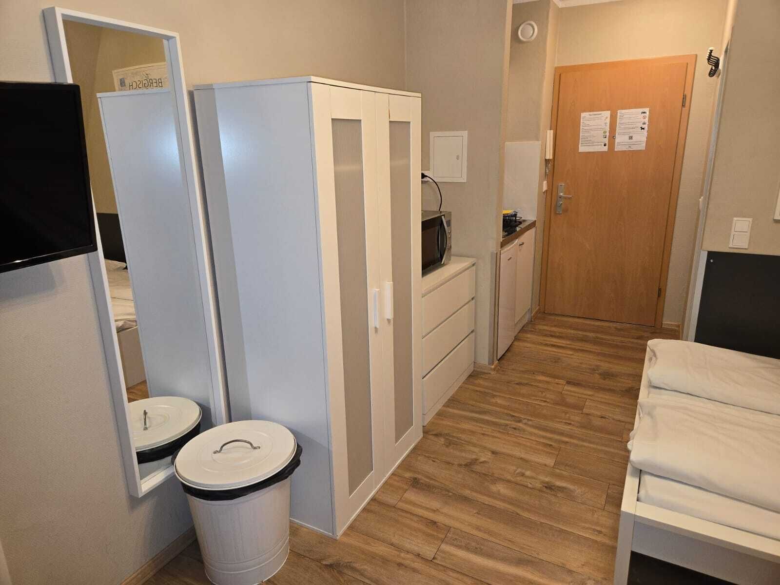 A compact hotel room with a bed, a small kitchenette, a mirror, a trash bin, and a wooden floor.