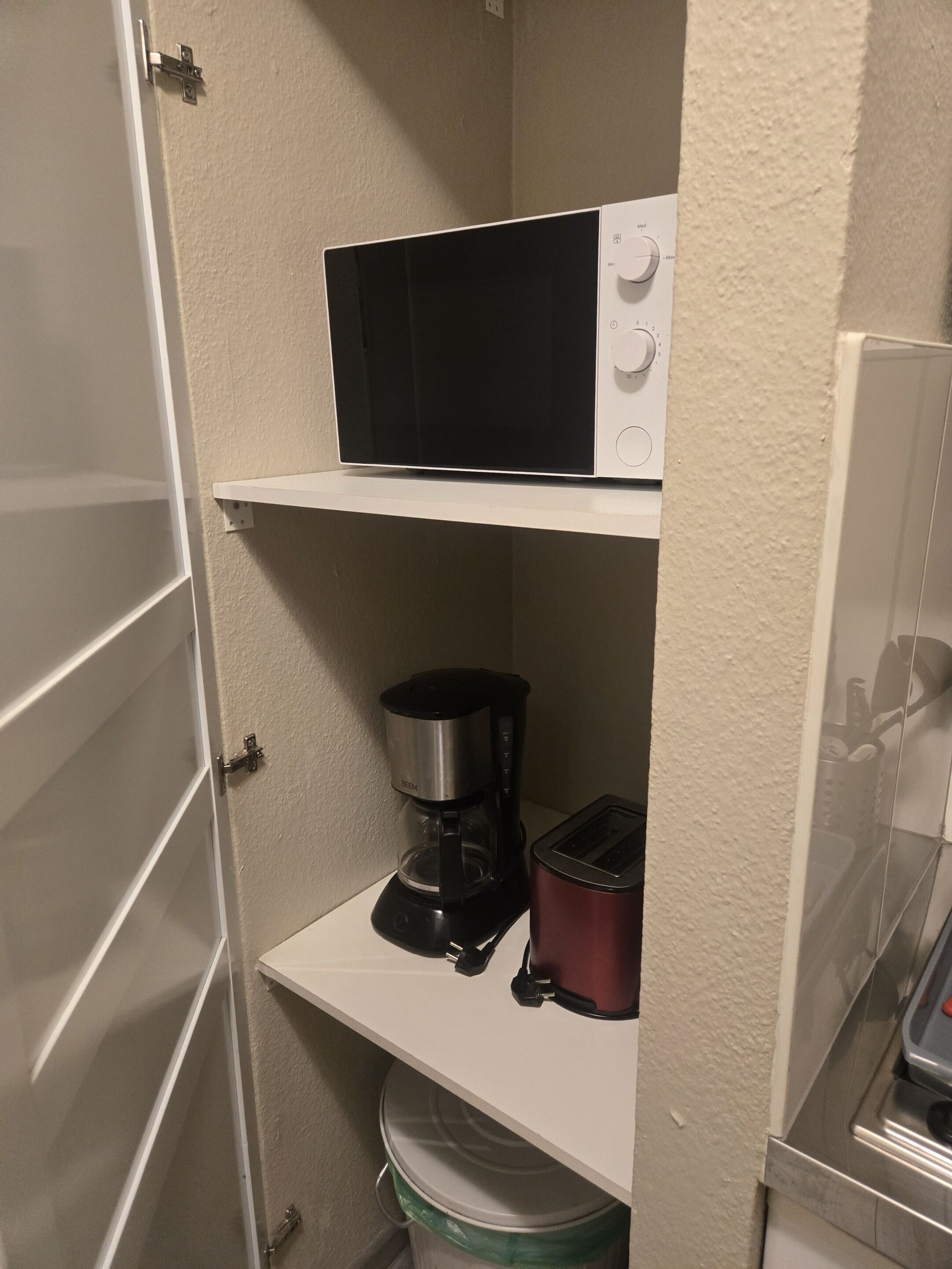 A compact kitchenette with a microwave, coffee maker, and toaster on white shelves.