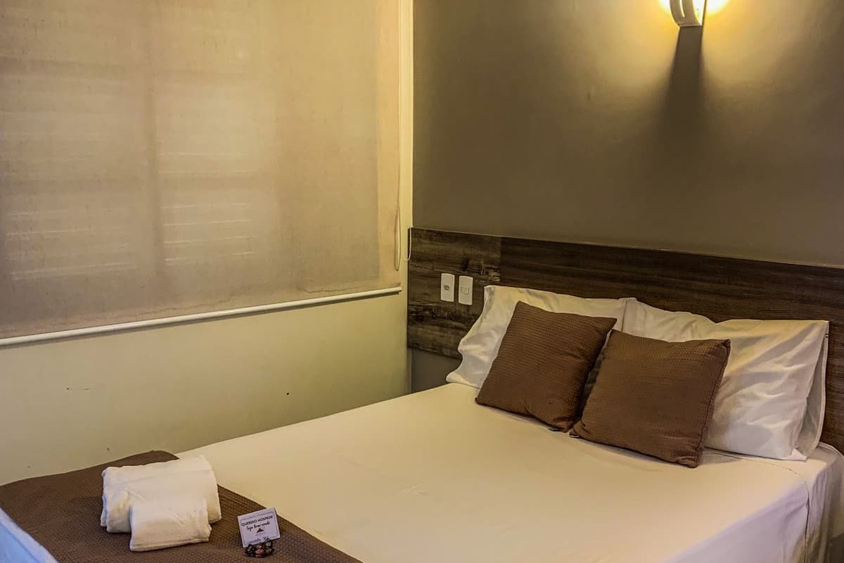 A hotel room with a bed, a bedside table, a wall-mounted air conditioner, and a wall-mounted light fixture.