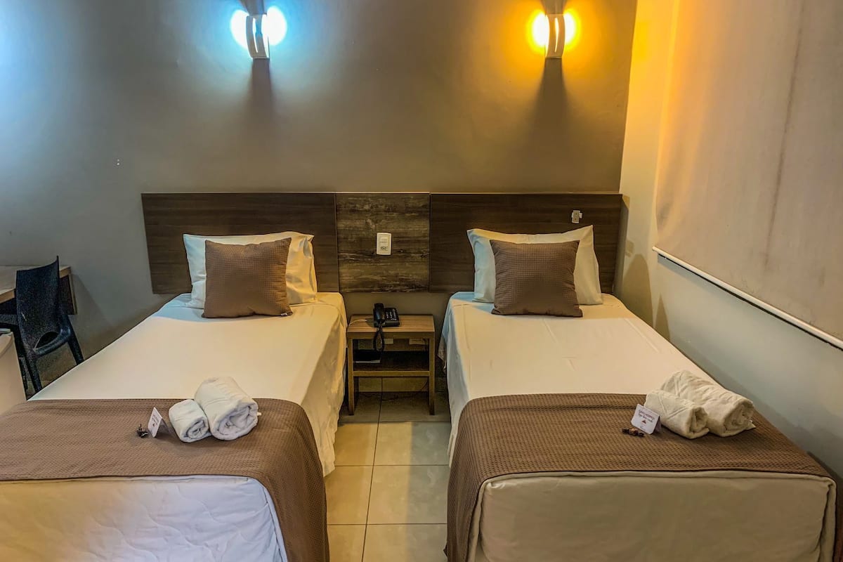 A hotel room with two beds, each with a brown bedspread and white linens. There are two bedside tables with lamps, a chair, and a small desk with a chair.