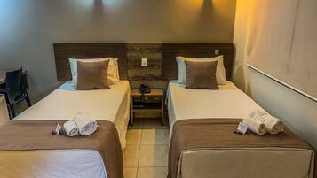 Quarto de hotel com duas camas, cada uma com colcha marrom e lençóis brancos. Há duas mesas de cabeceira com luminárias, uma cadeira e uma pequena escrivaninha com cadeira.