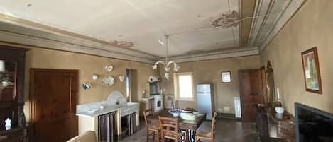 A spacious living room with a sofa, dining table, and chairs. There is a kitchen area with a refrigerator, stove, and cabinets. The room has a high ceiling with decorative moldings.
