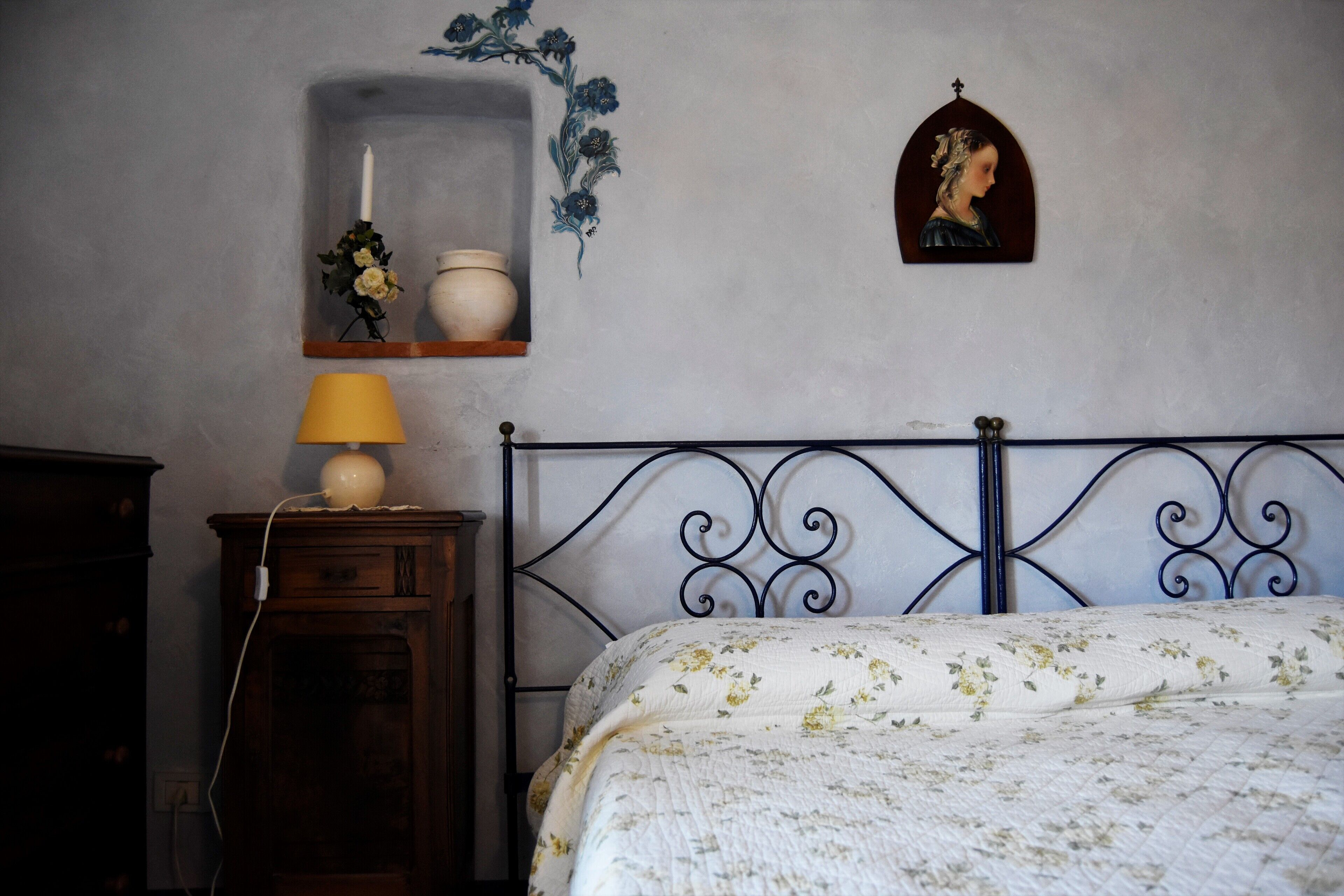 Una camera da letto con un letto, un comodino con una lampada, una mensola a parete con una candela e fiori, e un ritratto incorniciato.