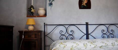 A bedroom with a bed, a bedside table with a lamp, a wall-mounted shelf with a candle and flowers, and a framed portrait.
