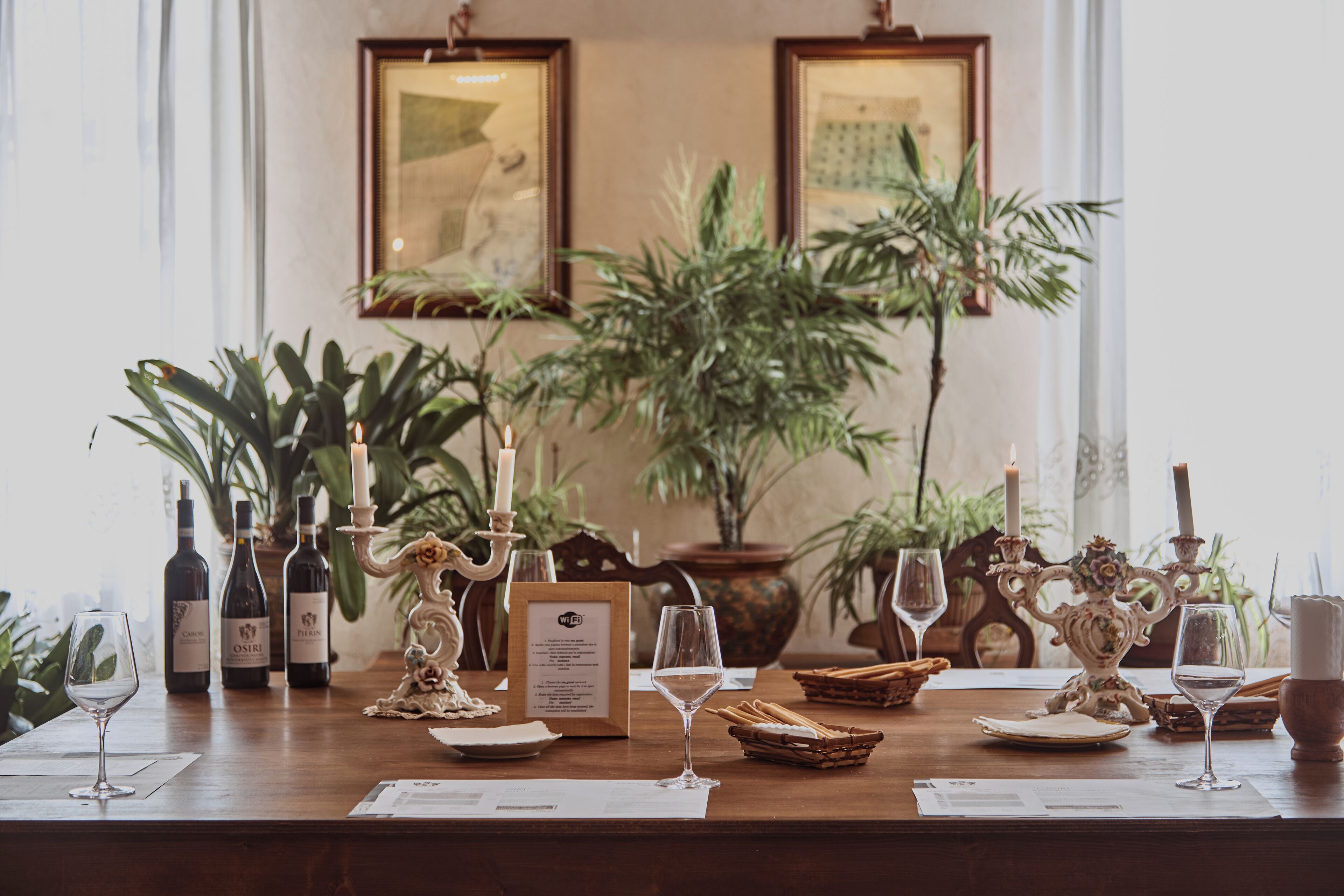 A dining table set with wine glasses, bottles, and candle holders, surrounded by potted plants and framed artwork.
