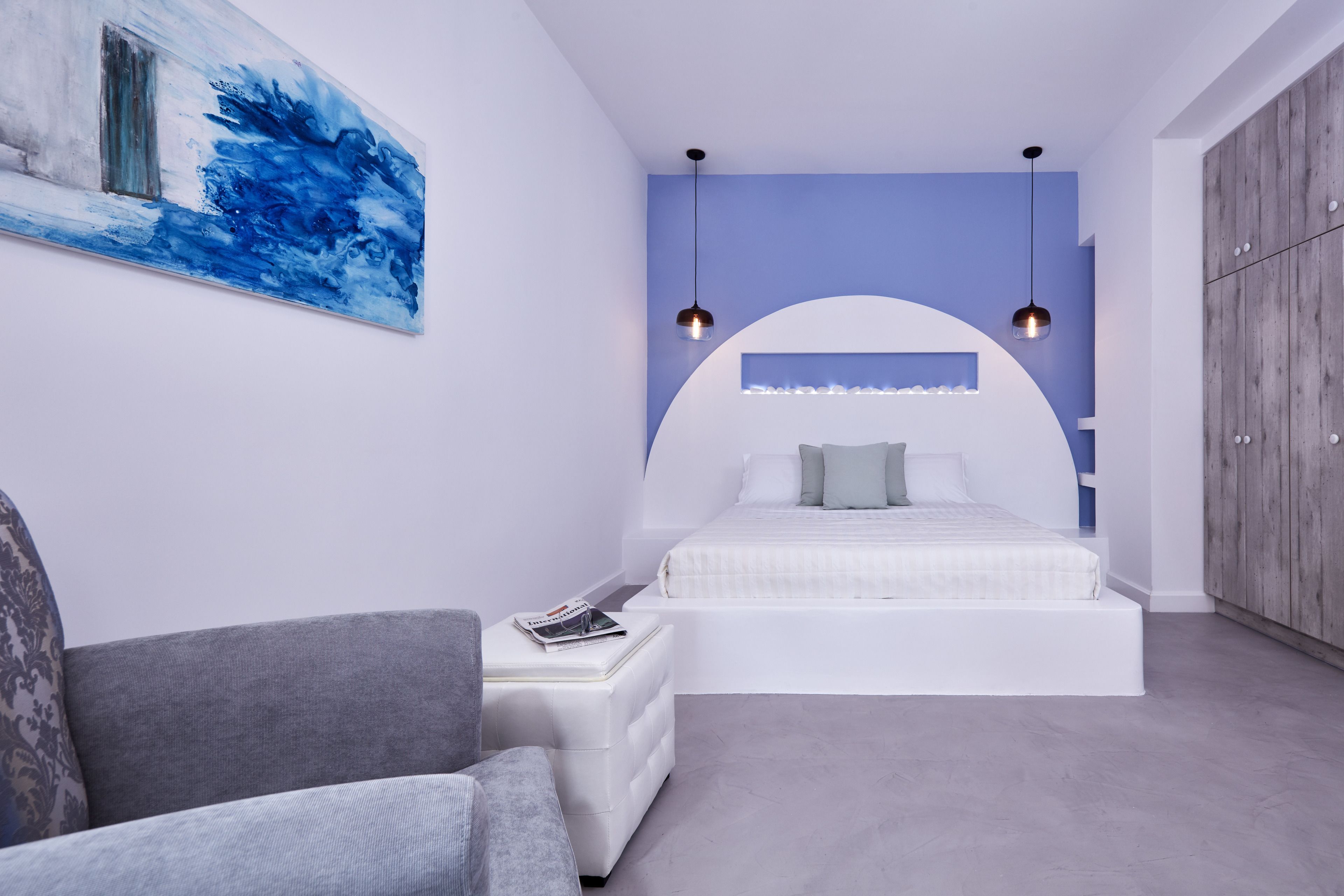 Un dormitorio moderno con una cama blanca curvada, un sofá gris y un cuadro abstracto azul en la pared.