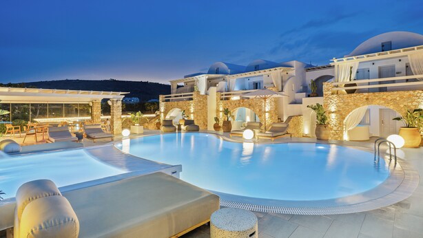 Piscina con sillones de descanso y una pared de piedra, rodeada de edificios blancos con cúpulas.