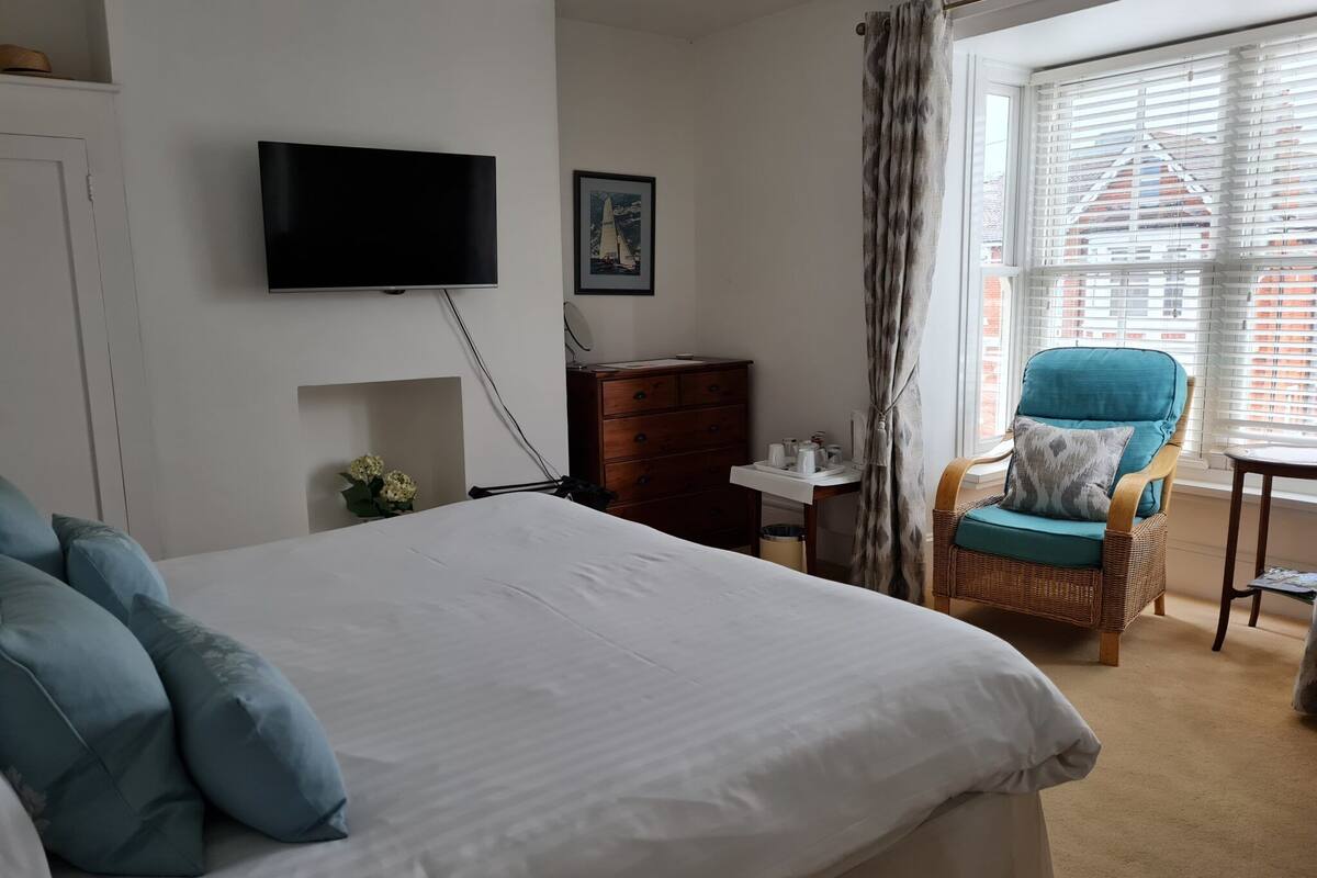 Une chambre à coucher comprenant un lit, un téléviseur fixé au mur, une commode, une chaise et une fenêtre avec des stores.