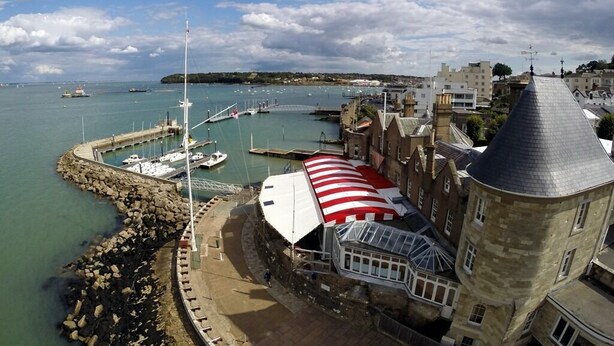 A coastal town with a prominent tower, a marina with boats, and a building with a striped awning. - Endeavour House (Cowes)