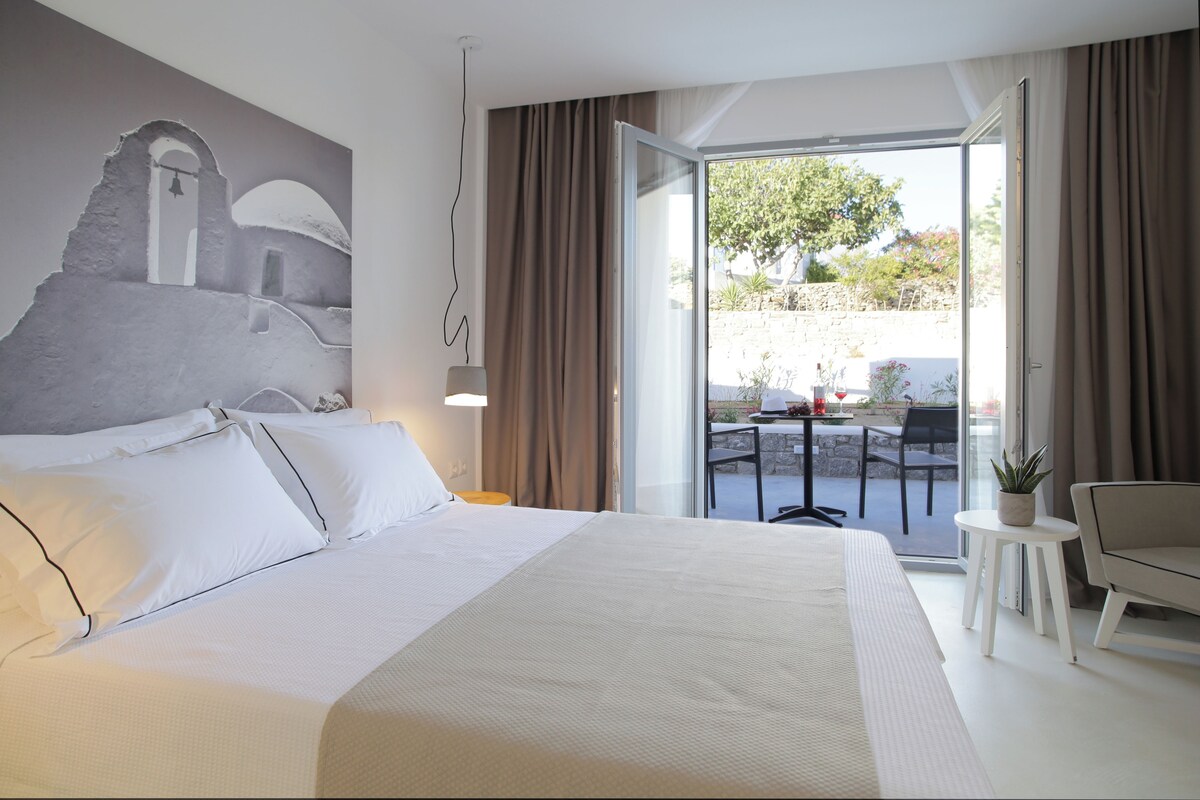 Une chambre d’hôtel avec un grand lit, un tableau représentant un bâtiment, un balcon avec une table et des chaises, et une plante en pot.