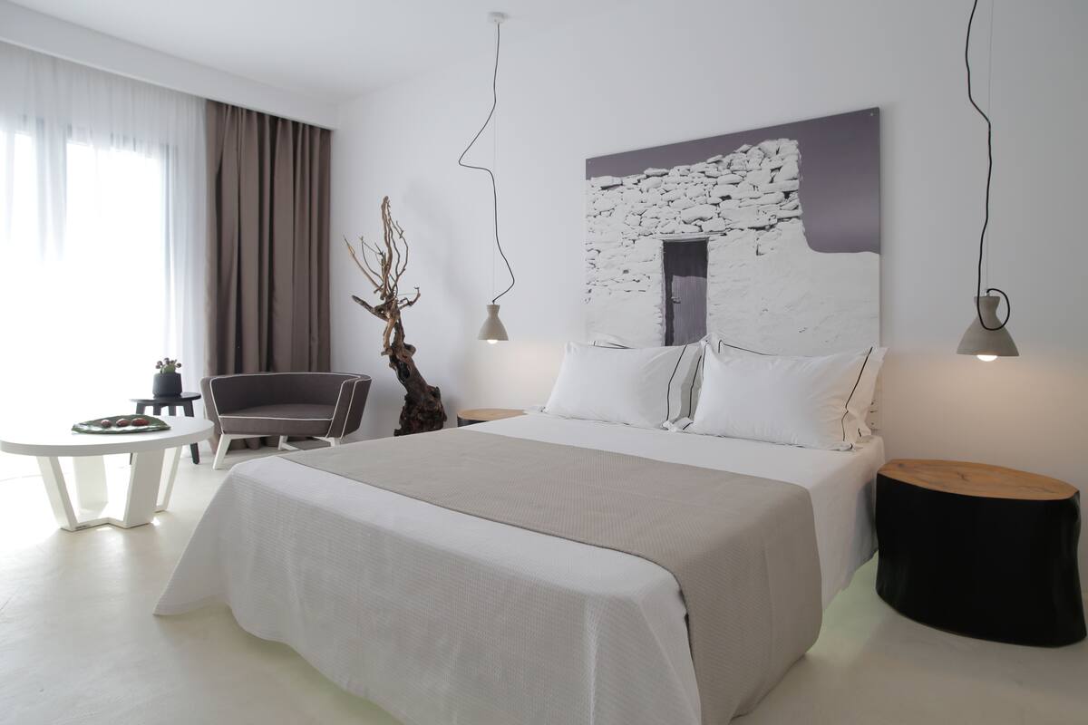 A modern hotel room with a large bed, a seating area with a chair and table, and a wall-mounted artwork.