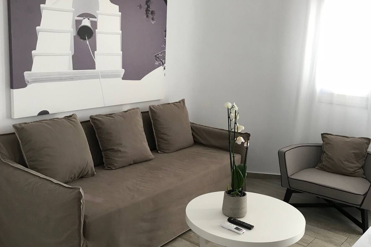 A modern living room with a brown sofa, a white coffee table, and a painting of a white church on the wall.