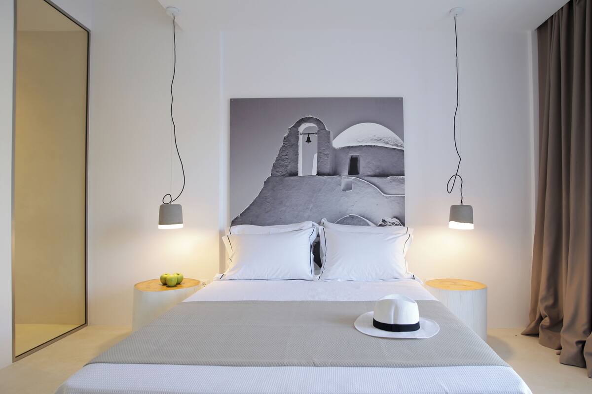 A modern hotel room with a bed, white bedding, a hat on the bed, a painting of a coastal scene, and two bedside lamps.