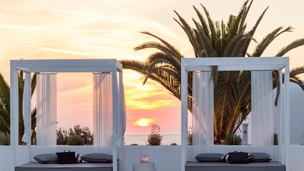 A serene poolside area with two white cabanas, each with a cushioned bench and a small table, set against a sunset backdrop. - Livin Mykonos Hotel - Adults Only by Mr and Mrs White (Mykonos)