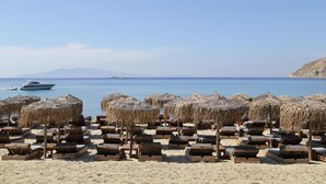 A beach with wooden lounge chairs and thatched umbrellas, a boat in the water, and a mountain in the background. - Livin Mykonos Hotel - Adults Only by Mr and Mrs White (Mykonos)
