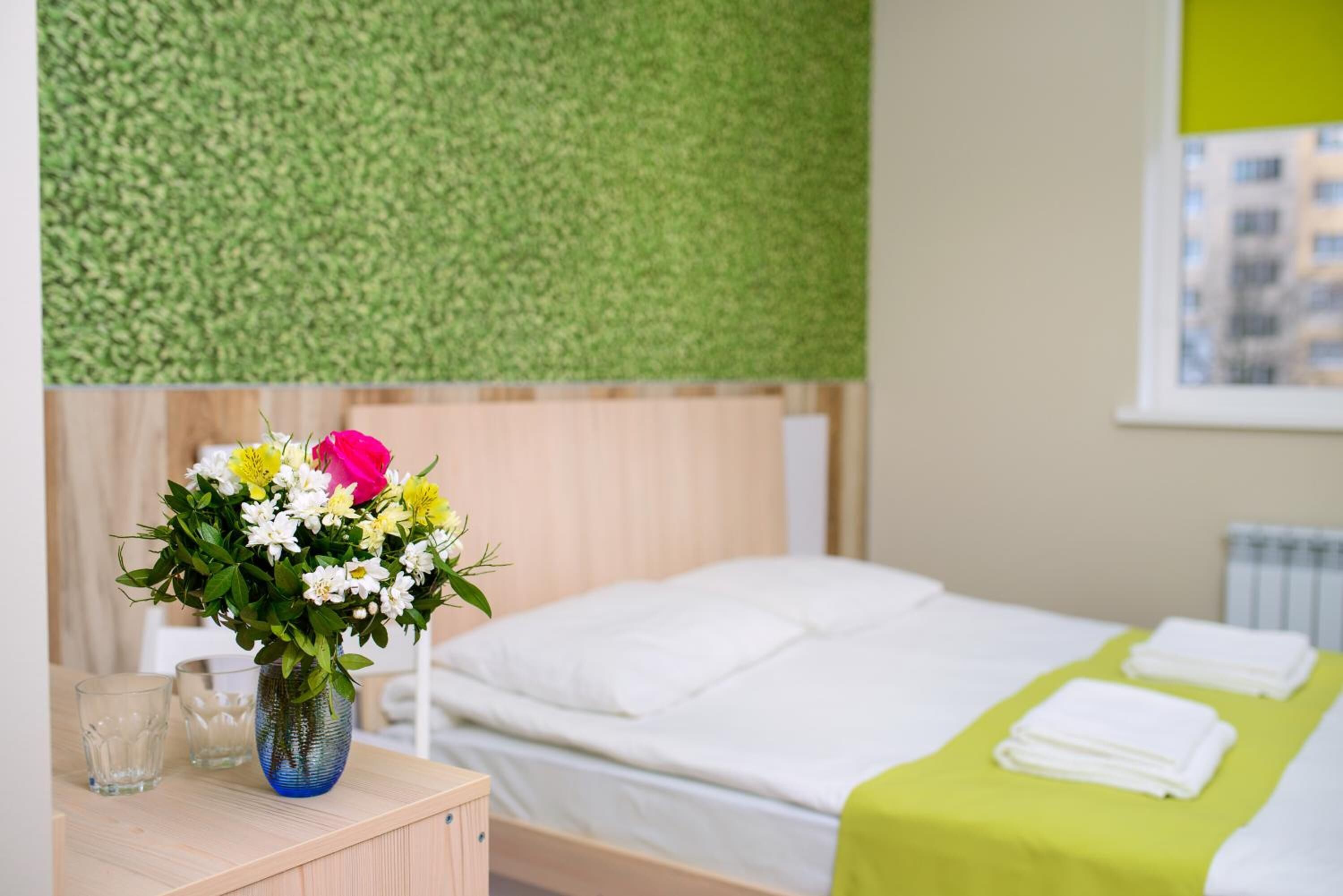 A hotel room with a bed, a nightstand with a vase of flowers, and a view of a building through a window.