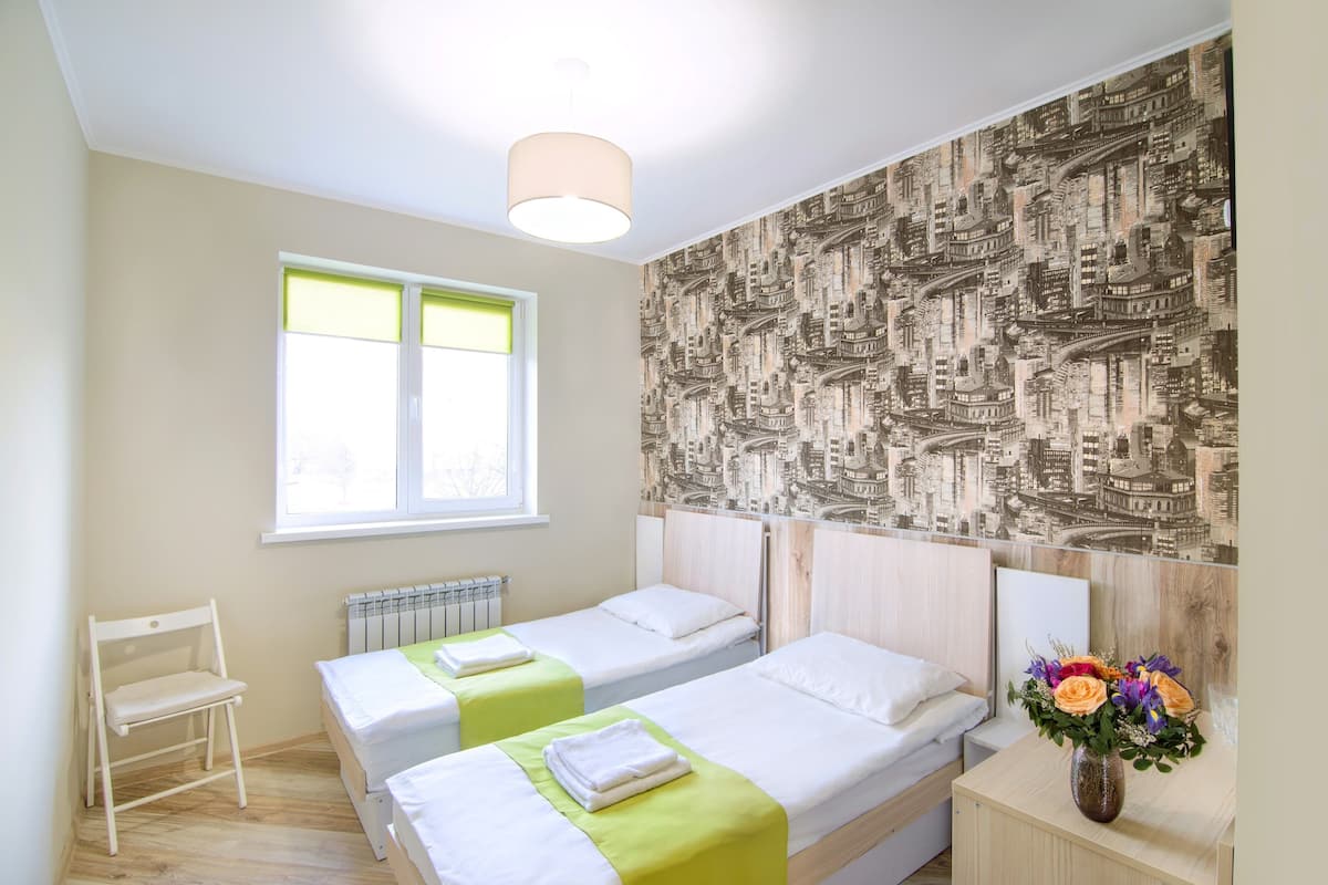 A hotel room with two beds, a radiator, a window with blinds, and a cityscape wallpaper.