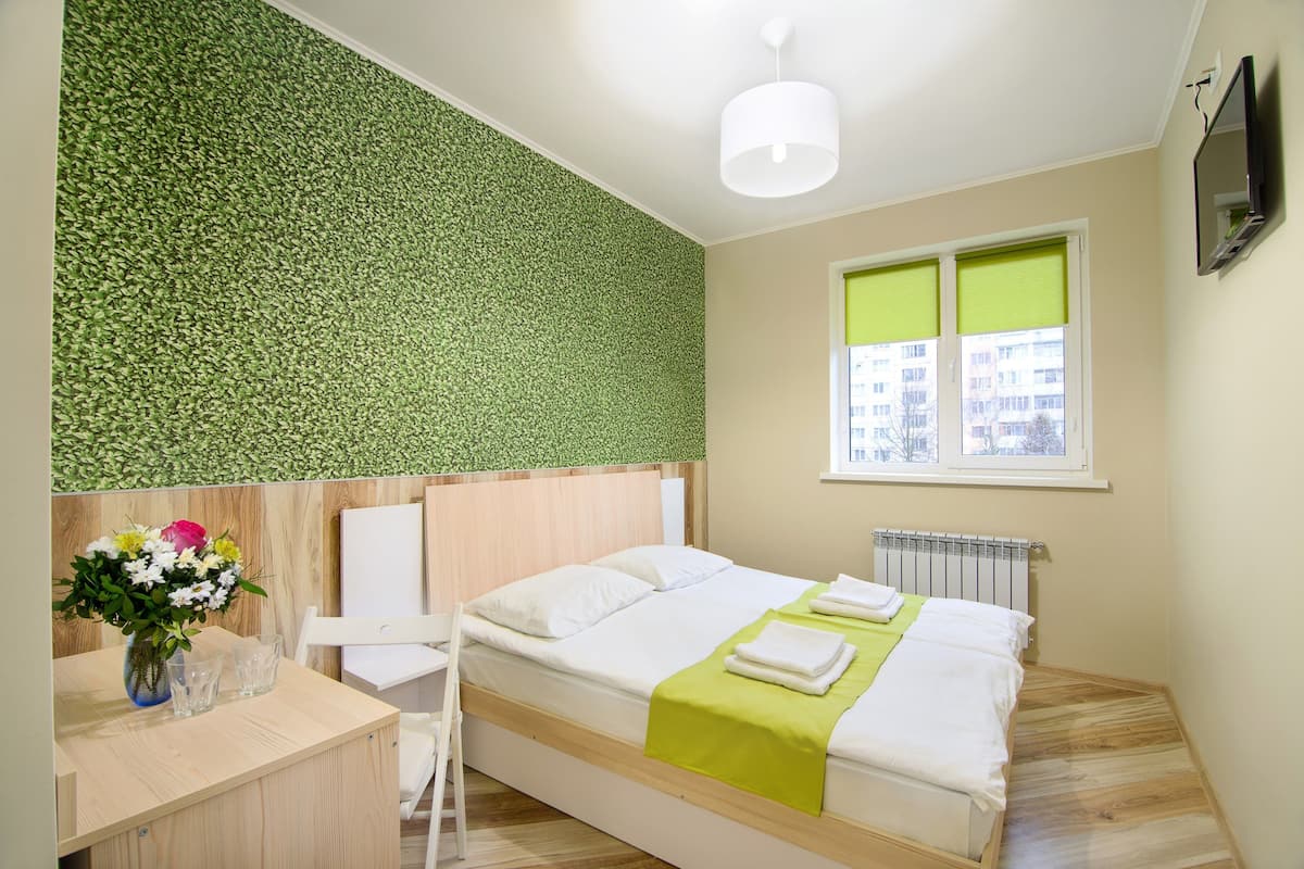 A bedroom with a bed, a desk, a chair, a window with blinds, and a radiator.