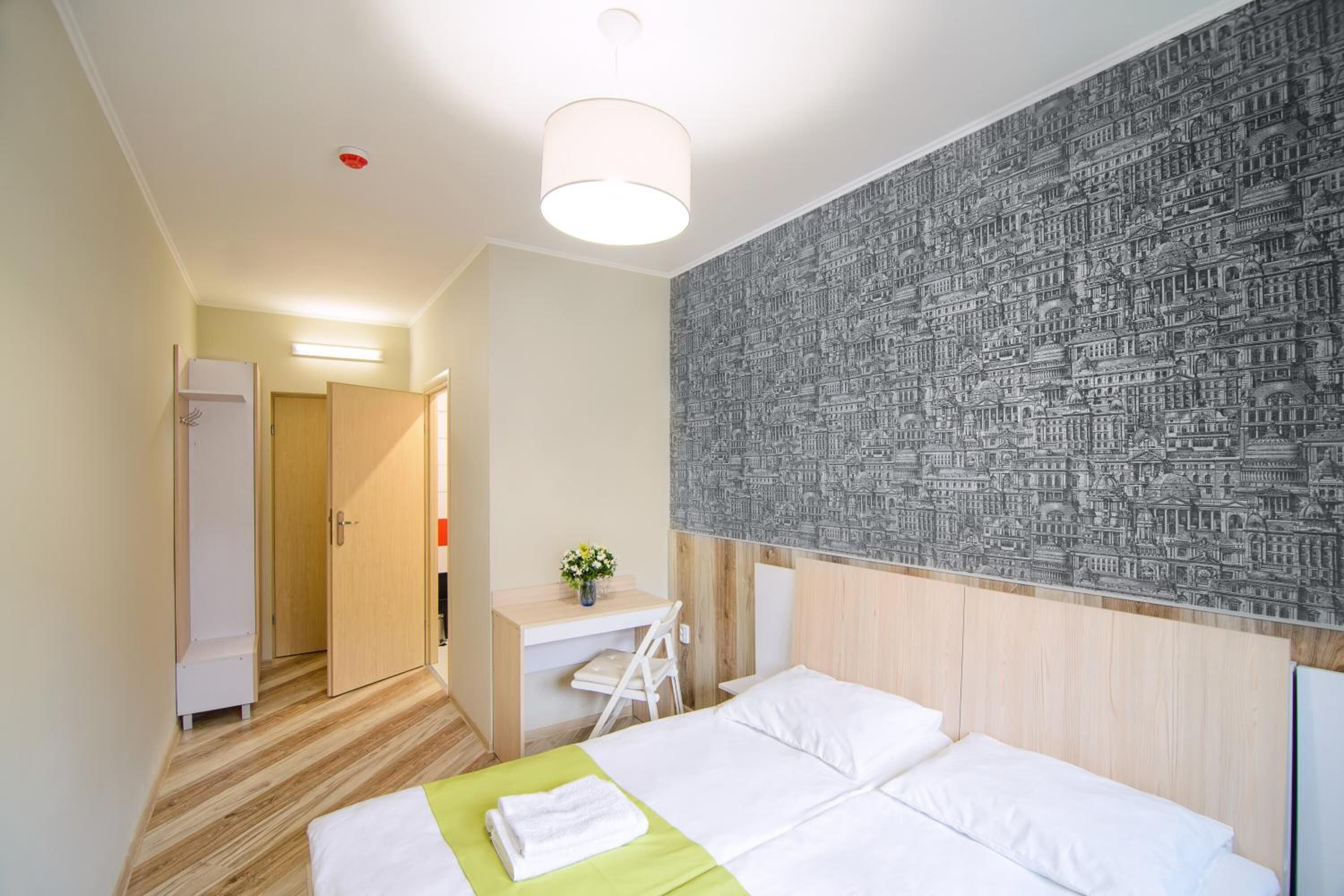 A hotel room with a bed, a desk, a chair, and a wall with a cityscape wallpaper.