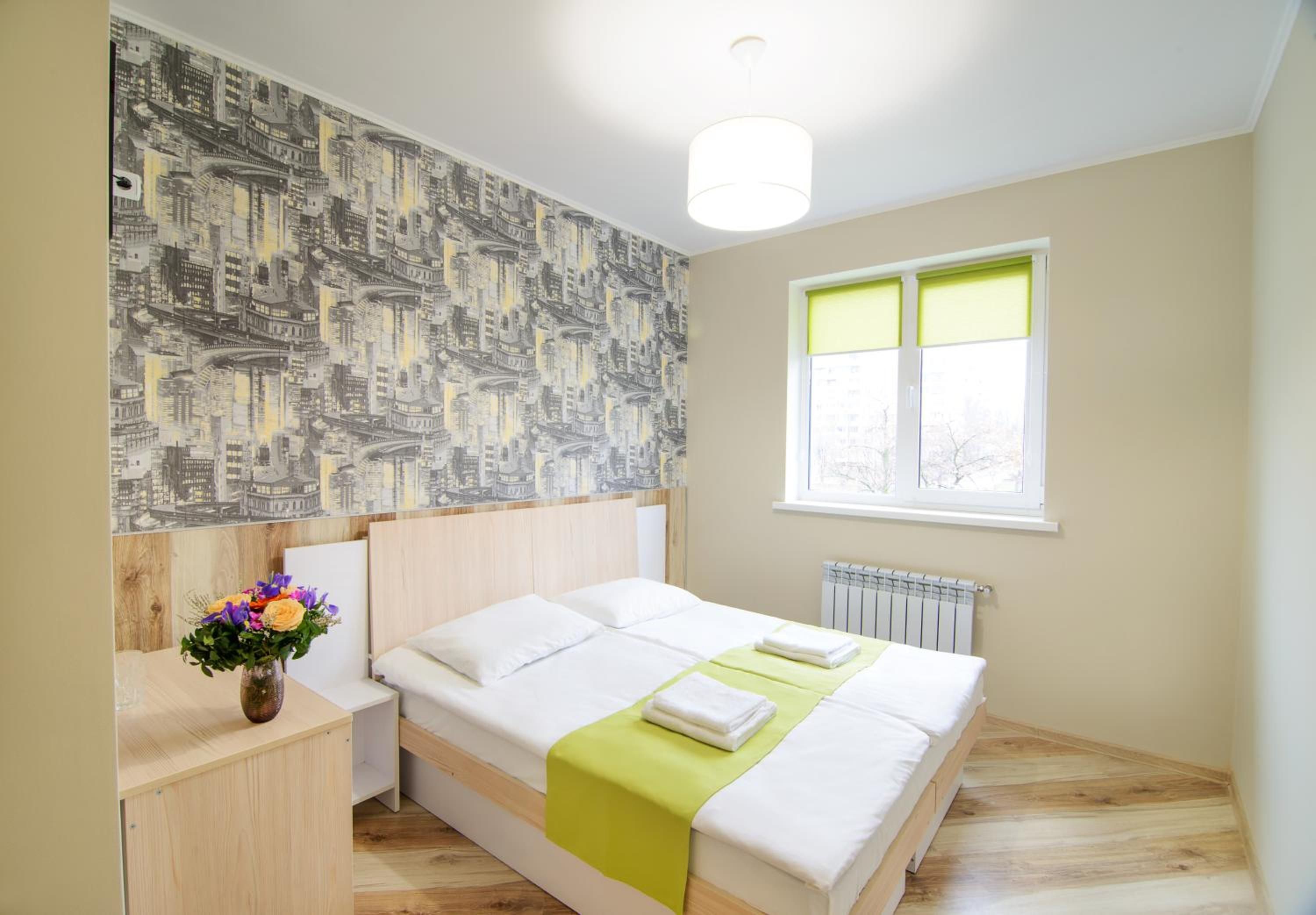 A hotel room with a bed, a nightstand, a vase with flowers, a window with blinds, and a radiator.