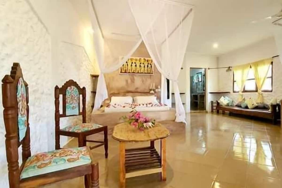 A spacious room with a large bed, a wooden chair, a small table with flowers, and a sofa.
