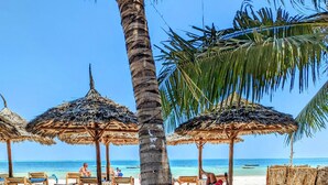 Ein Essbereich am Strand mit Holztischen und -stühlen, Reetdachschirmen und einer Palme.