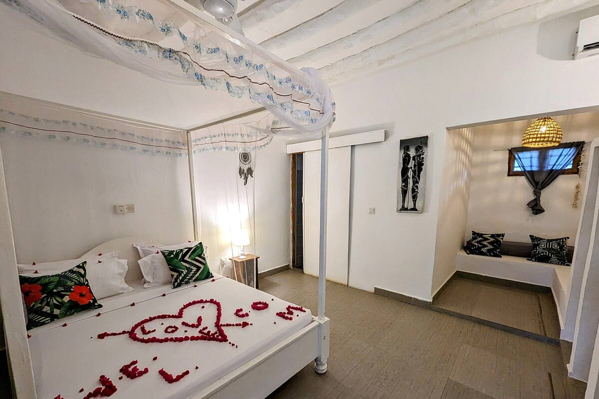 A four-poster bed with rose petals arranged on the bedspread, a bedside lamp, and a wall-mounted artwork.