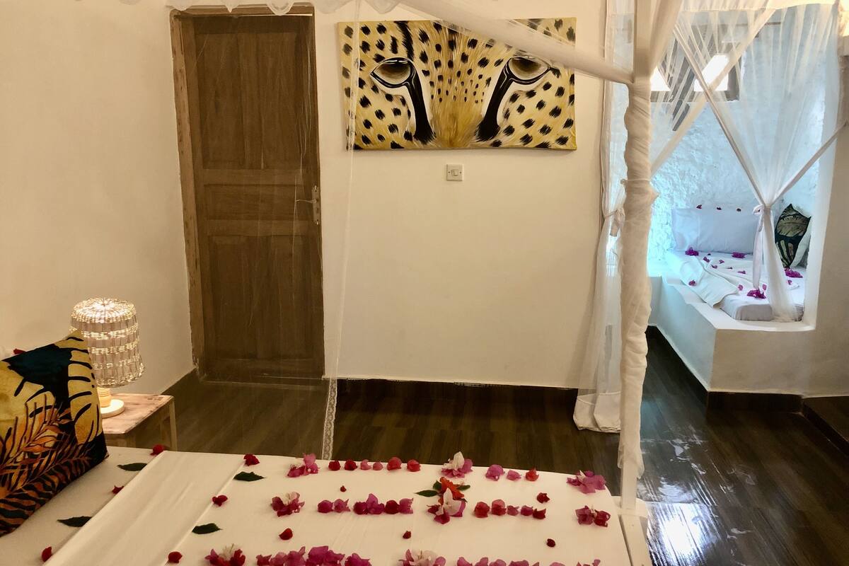 A room with a bed adorned with rose petals, a wooden door, a painting on the wall, and a window with a white curtain.