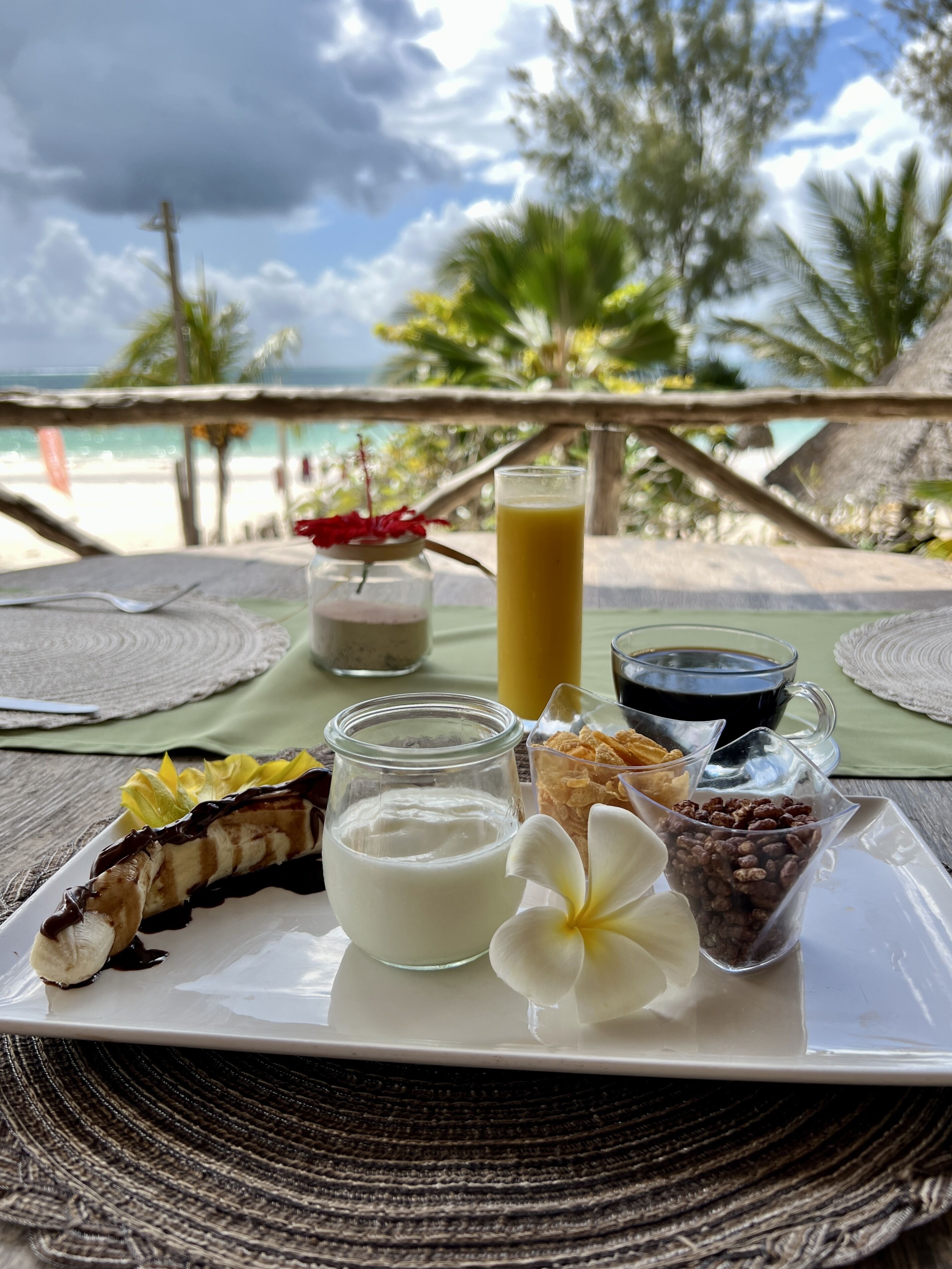 A breakfast tray with various food items and drinks on a table with a view of the beach and palm trees.