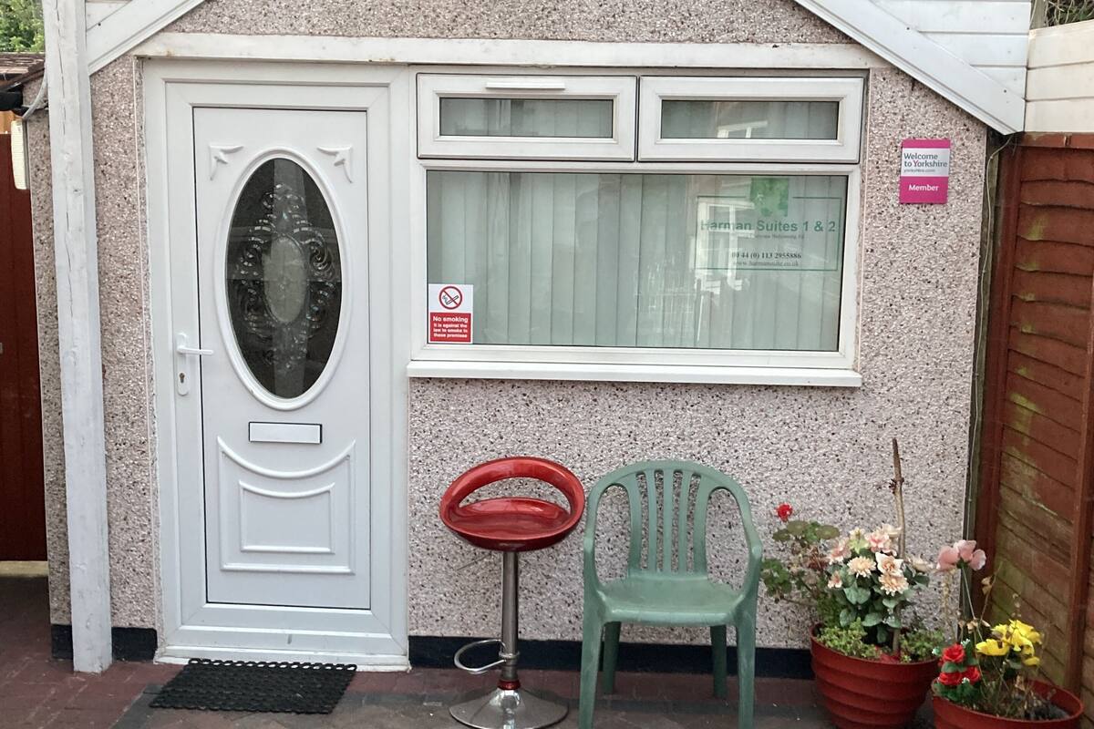 A small building with a white door, a window with a sign, a green chair, and potted plants.