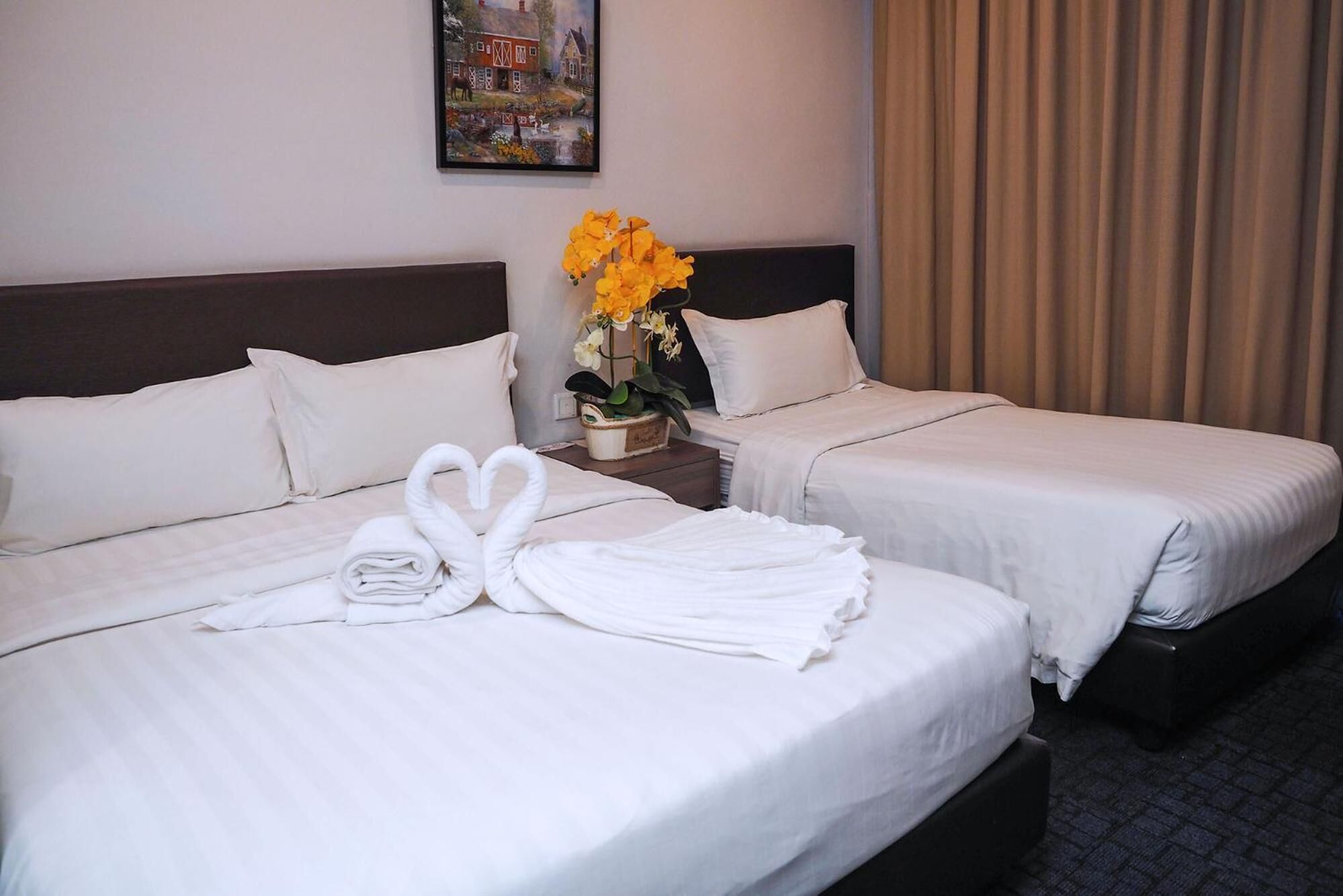 A hotel room with two beds, white linens, a heart-shaped towel arrangement, a vase with yellow flowers, and a painting on the wall.