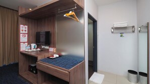 Quarto de hotel com cama de madeira, televisão, pequena escrivaninha com bandeja de produtos de higiene e toalheiro, e lixeira.