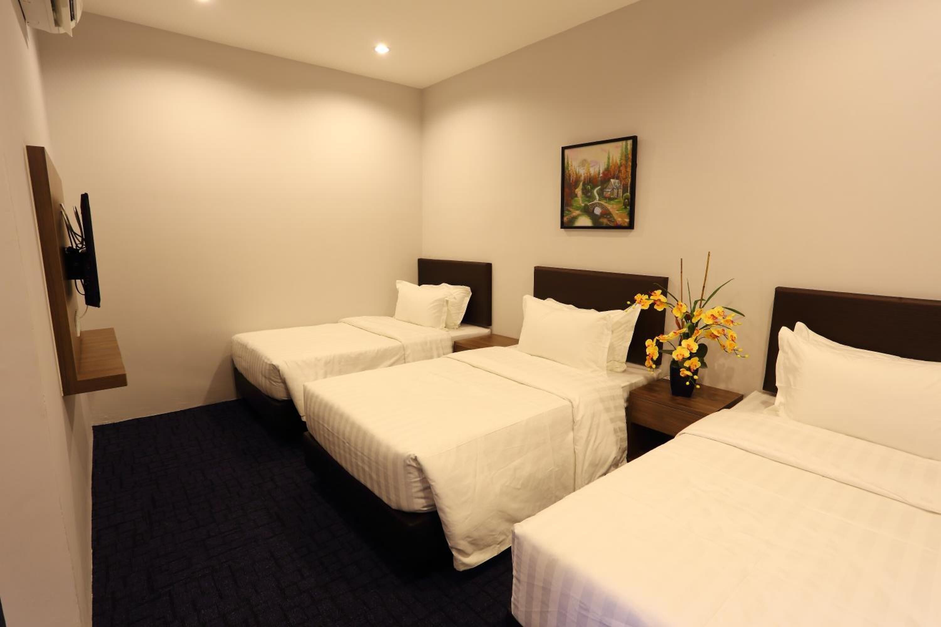 A hotel room with three single beds, a small table with a vase of flowers, and a painting on the wall.